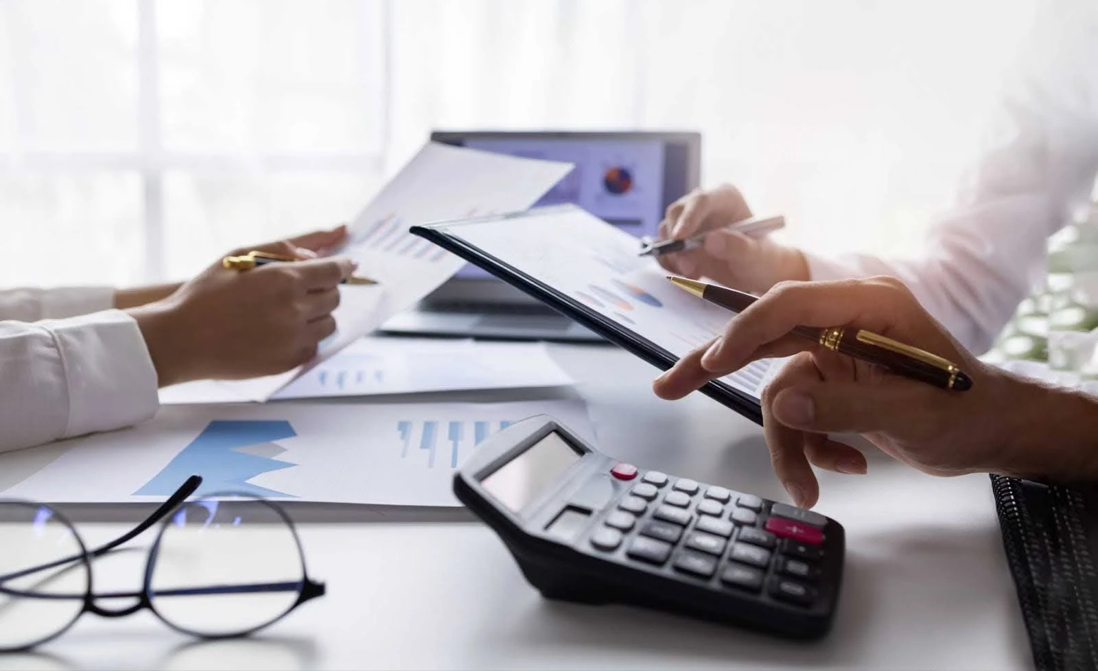Imagem que retrata pessoas analisando gráficos e relatórios financeiros em um ambiente de trabalho, com uma calculadora ao fundo, ideal para temas de finanças.