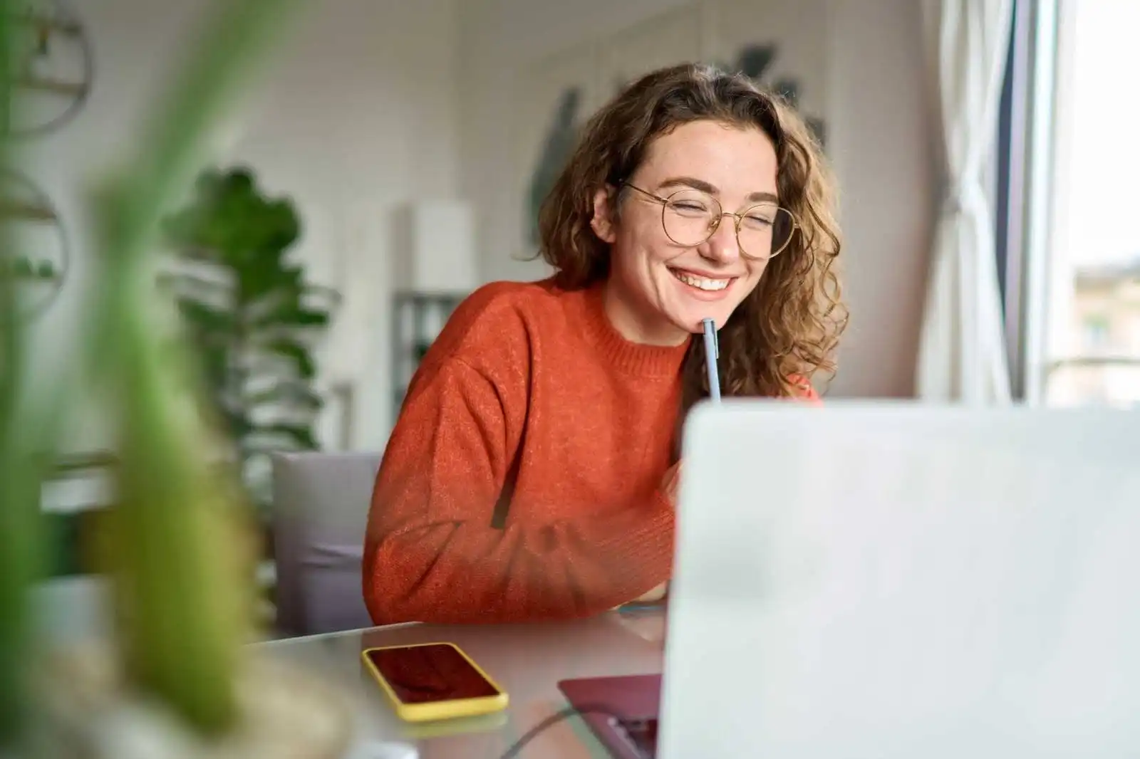 Mulher feliz trabalhando em um laptop, com caneta na mão e usando óculos, em ambiente aconchegante e iluminado. Ótima imagem para temas de produtividade e home office.
