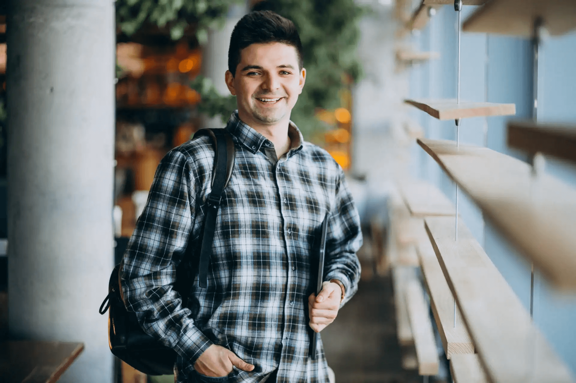 Homem sorridente usando camisa xadrez e mochila, fazendo compras em loja de móveis ou decoração, com prateleiras de madeira ao lado e ambiente iluminado.
