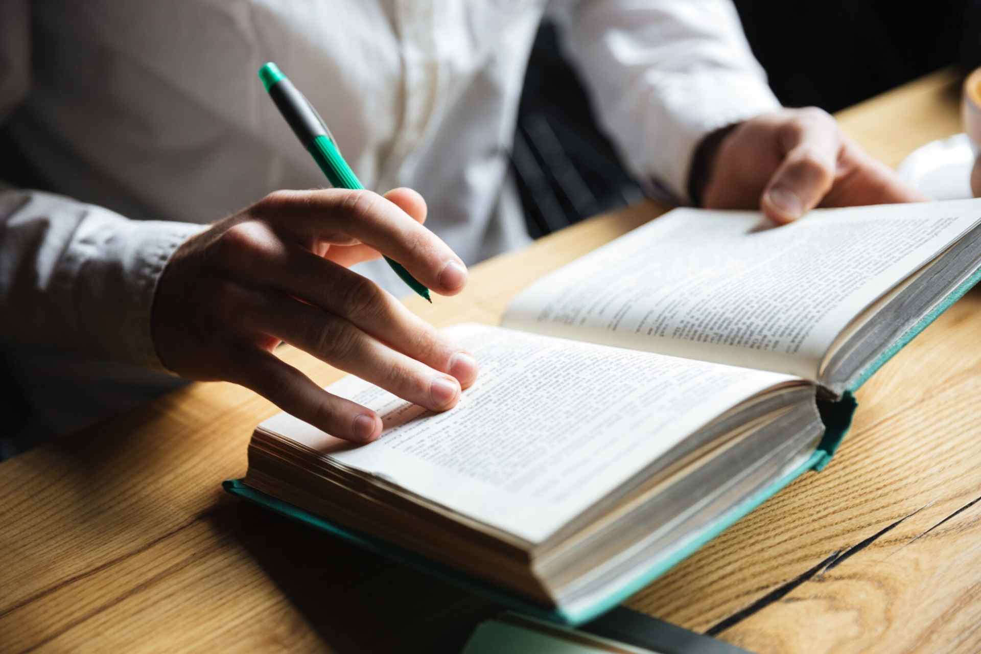 Pessoa segurando um marcador verde e escrevendo em um livro aberto sobre uma mesa de madeira, com foco na leitura e anotações durante o estudo.