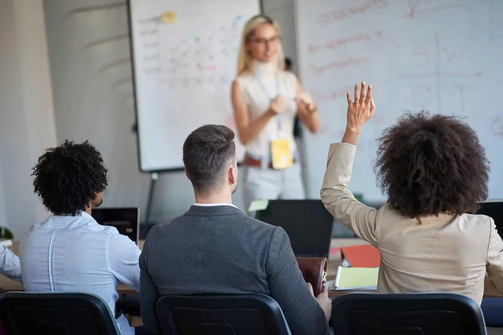 Aula ativa em um ambiente de negócios, onde uma mulher apresenta enquanto os alunos participam ativamente, levantando a mão para fazer perguntas.