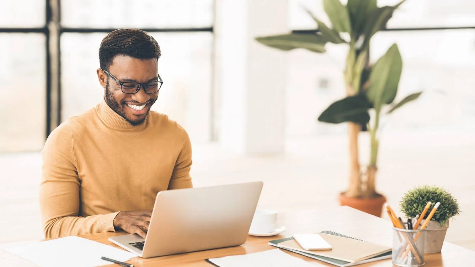 Homem sorridente trabalhando em notebook em um ambiente iluminado. Escritório moderno com plantas e material de escritório. Trabalho remoto produtivo.