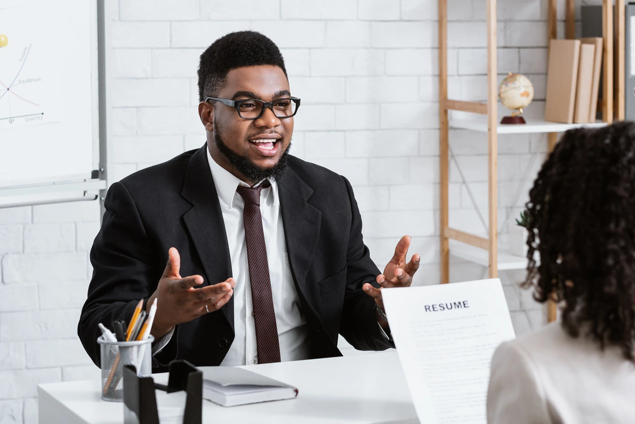 Entrevista de emprego com um homem em terno conversando com uma mulher, mostrando um currículo em um ambiente de escritório.