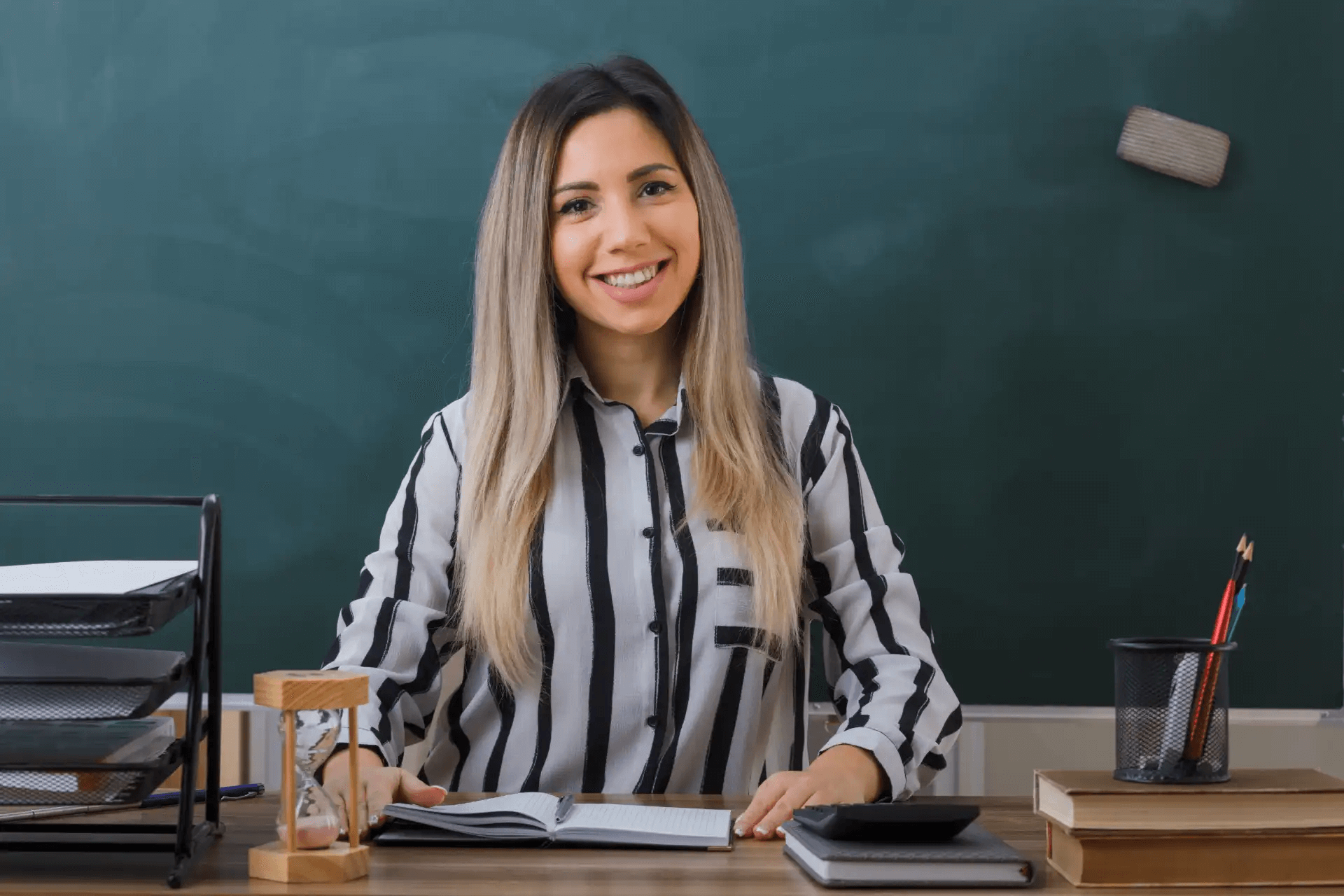 Professora sorridente em sala de aula, revisando materiais escolares e ensinando estudantes com entusiasmo, em uma sala com quadro-negro ao fundo.