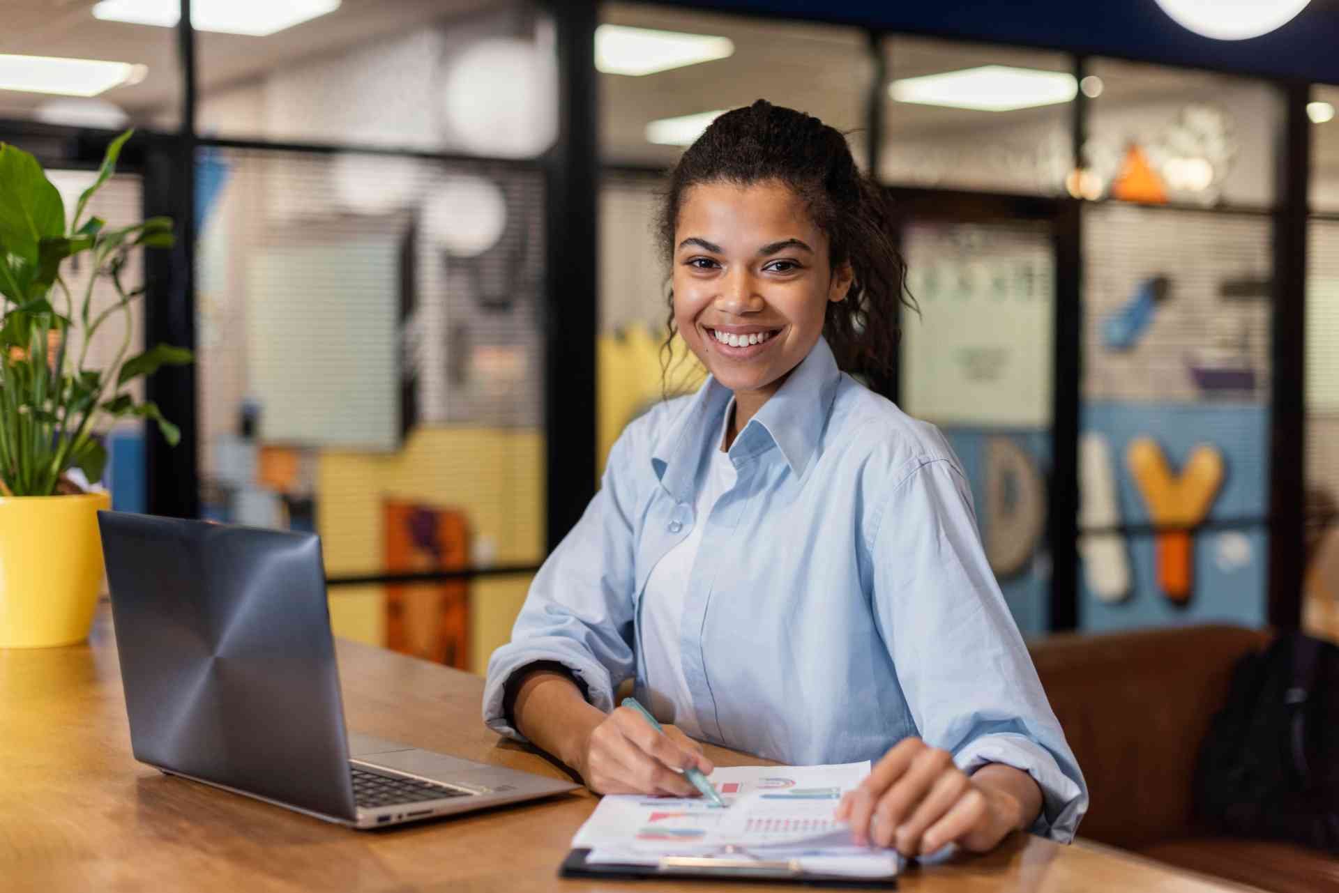 Atendente escolar sorridente sentado em mesa com laptop e caderno, trabalhando em ambiente escolar bem iluminado e organizado.