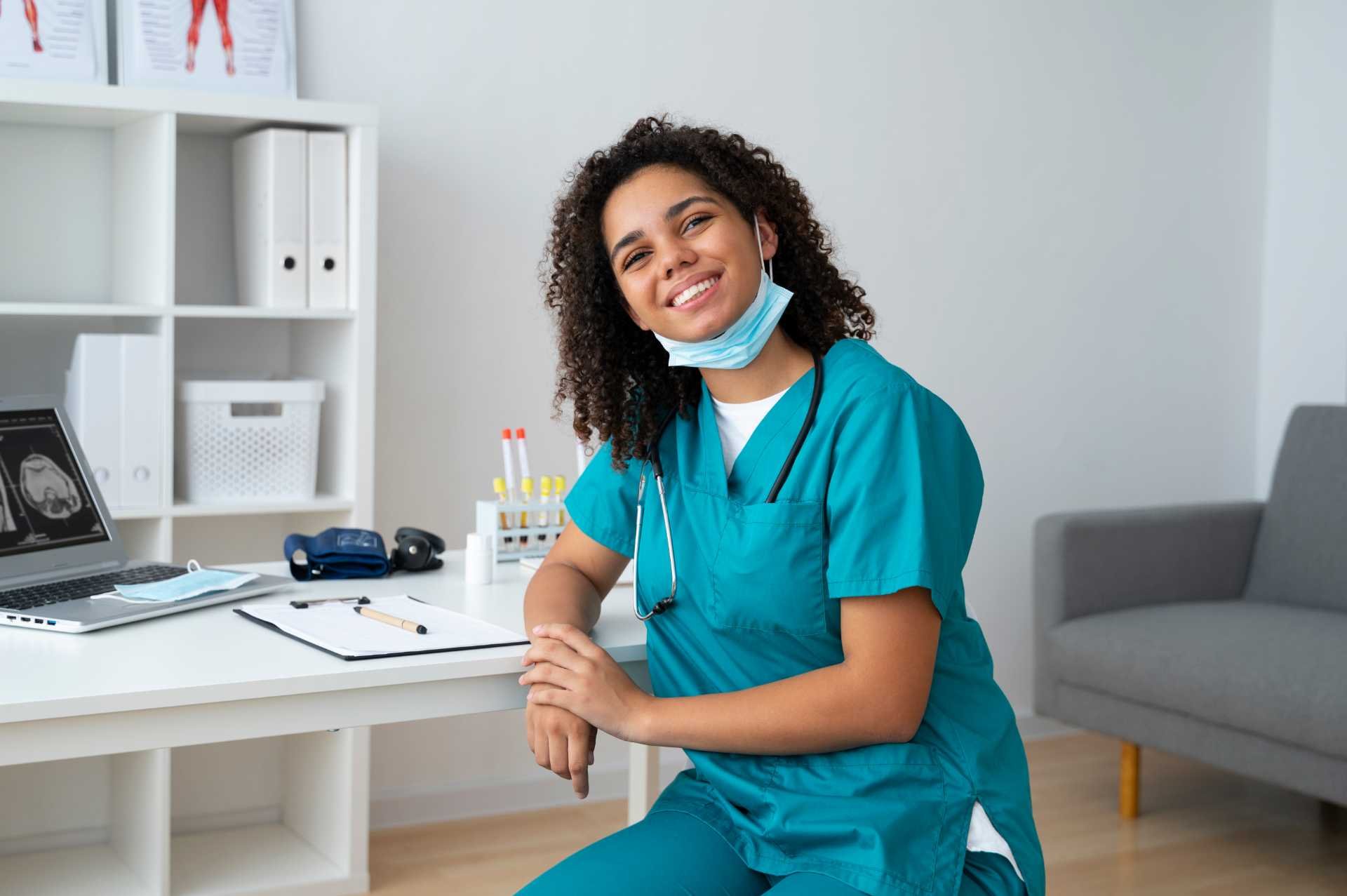 Enfermeira sorridente em consultório realizando atendimento de enfermagem do trabalho, com equipamentos e computador ao fundo.