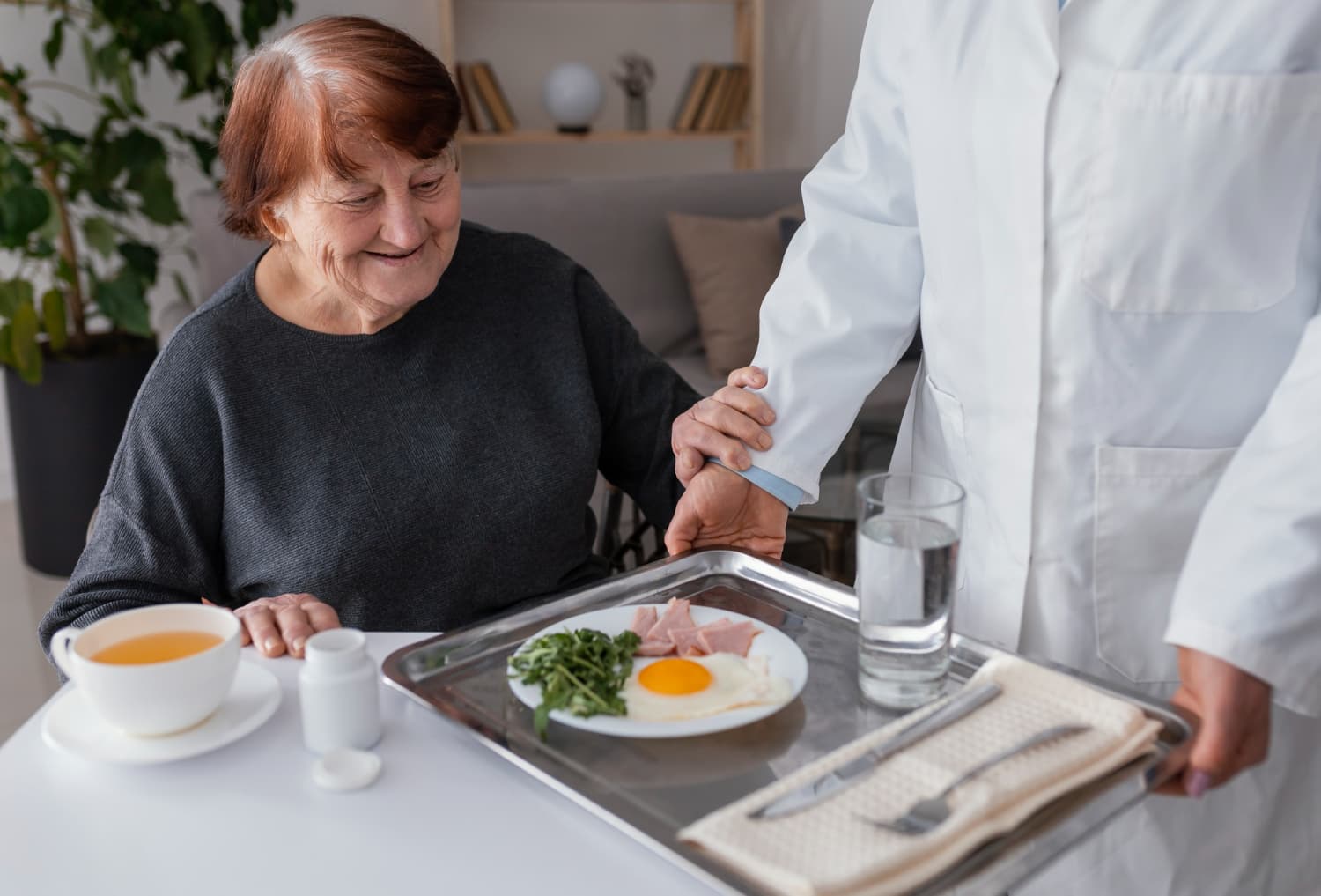 Mulher idosa sorrindo recebendo café da manhã na cama de um cuidador ou familiar, com prato de ovos, verduras, e Chá branco ao lado.