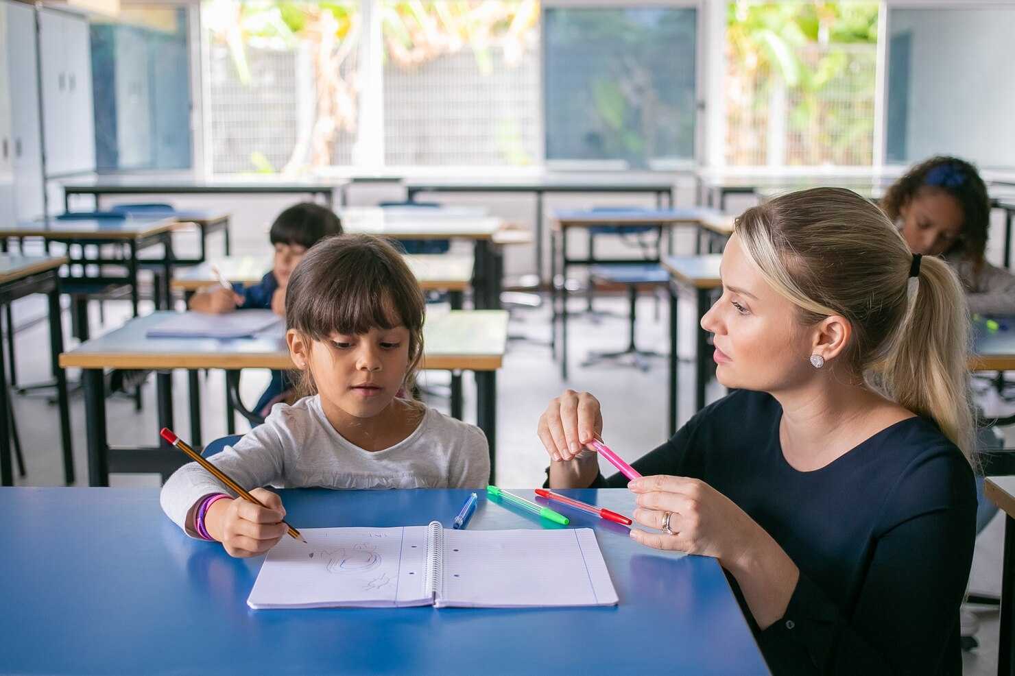 Professora ajudando uma criança a fazer tarefas escolares em sala de aula, com várias crianças ao fundo em um ambiente bem iluminado.