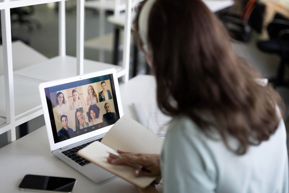 Pessoa em home office usando notebook para participar de videoconferência, com tela exibindo várias pessoas em chamada e anotações na mesa; ambiente de trabalho com celular ao lado.