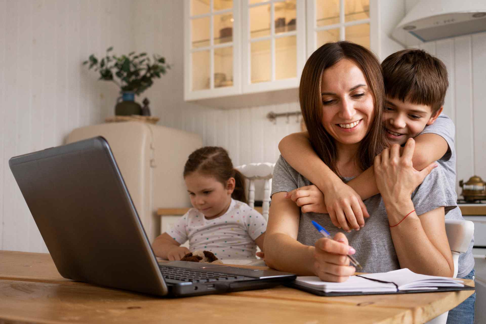 Retrato de mãe em casa conciliar trabalho e estudos enquanto cuida dos filhos: ela sorri abraçando a criança e usa um tablet, com caderno e lápis sobre a mesa.