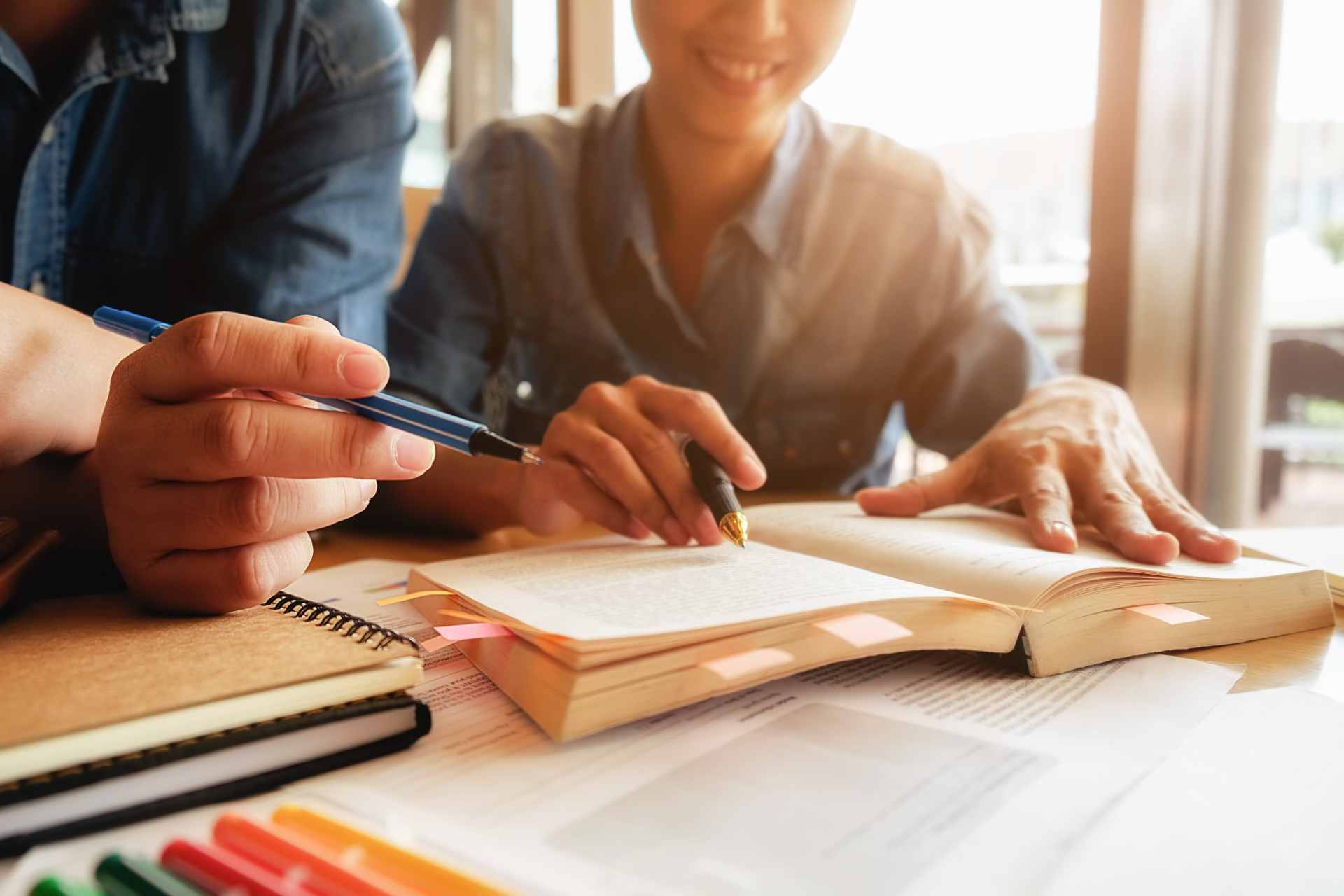 Pessoas estudando em grupo com livros e anotações na mesa, enquanto uma pessoa escreve com caneta durante cursos livres para desenvolver novas habilidades e crescimento profissional.