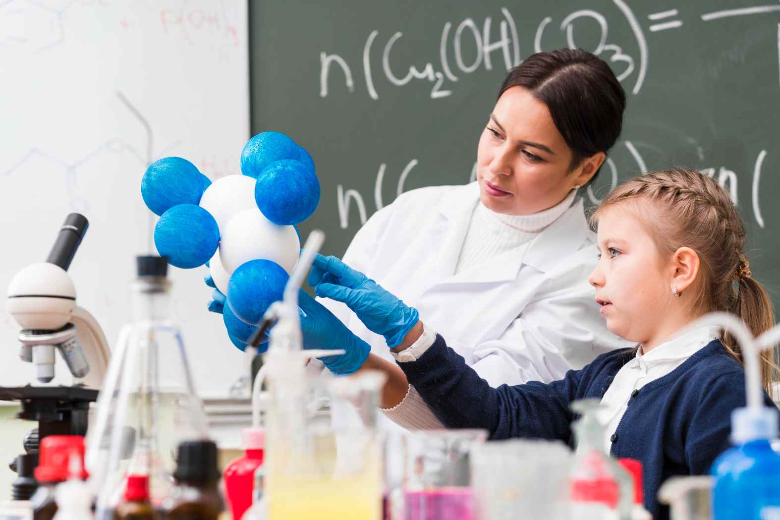 Professor(a) de Química em sala de aula orientando estudantes em experimento com esferas de moléculas e vidrarias, estimulando a aprendizagem científica.