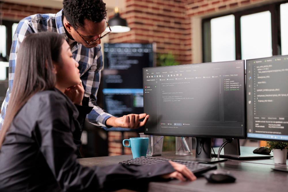 Duas pessoas em ambiente de escritório colaboram no desenvolvimento de software, analisando códigos na tela de um monitor e apontando para o computador, com xícara sobre a mesa.