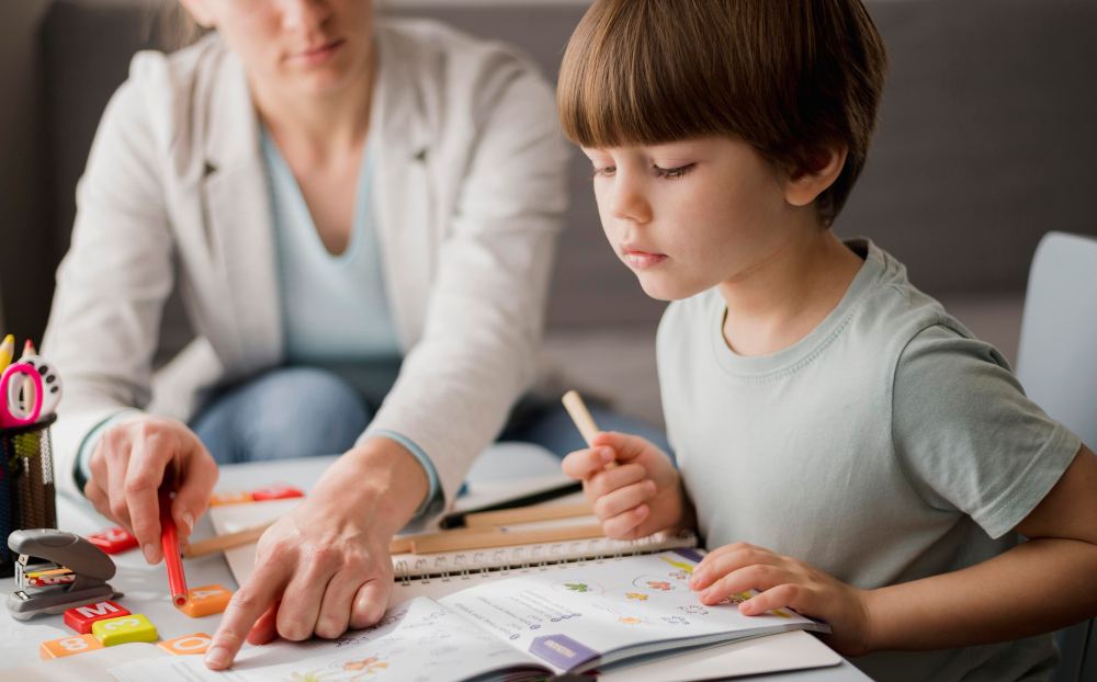 Criança e adulto estudando juntos em casa: o menino desenha e faz anotações em um caderno aberto enquanto a pessoa ao fundo acompanha a atividade com um sorriso. Foto realista.