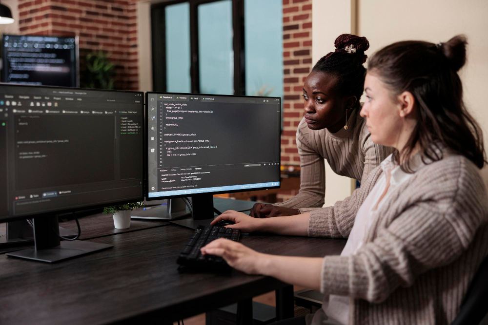 Duas pessoas trabalhando em computadores com monitores exibindo código de programação em um ambiente de escritório com parede de tijolos e plantas sobre a mesa.