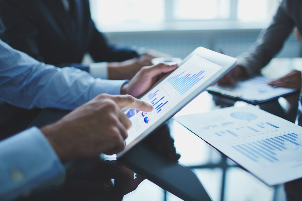 Equipe em reunião analisa relatórios e dados em tablets, com gráficos e tabelas na tela, sugerindo planejamento e tomada de decisão no ambiente corporativo.