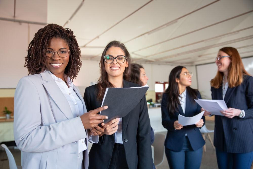 Equipe de mulheres profissionais em um ambiente corporativo, sorrindo e segurando pastas e documentos, com uma pessoa em destaque na frente, durante um trabalho ou reunião.