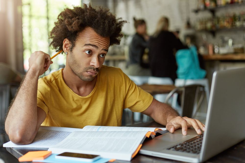 Homem jovem com cabelo afro usando o computador em casa, enquanto observa um papel e parece concentrado em um projeto ou trabalho.