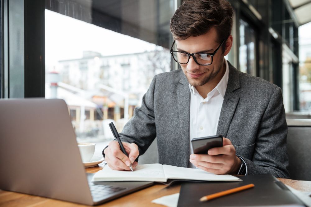 Homem de terno e óculos em ambiente corporativo, anotando em um caderno e usando um smartphone ao lado de um notebook