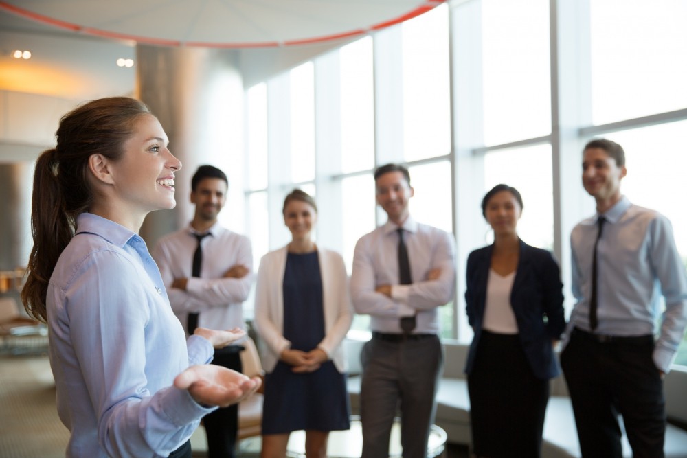Mulher apresentando em um ambiente corporativo moderno para um grupo de profissionais ao fundo, com clima de reunião e comunicação no escritório.