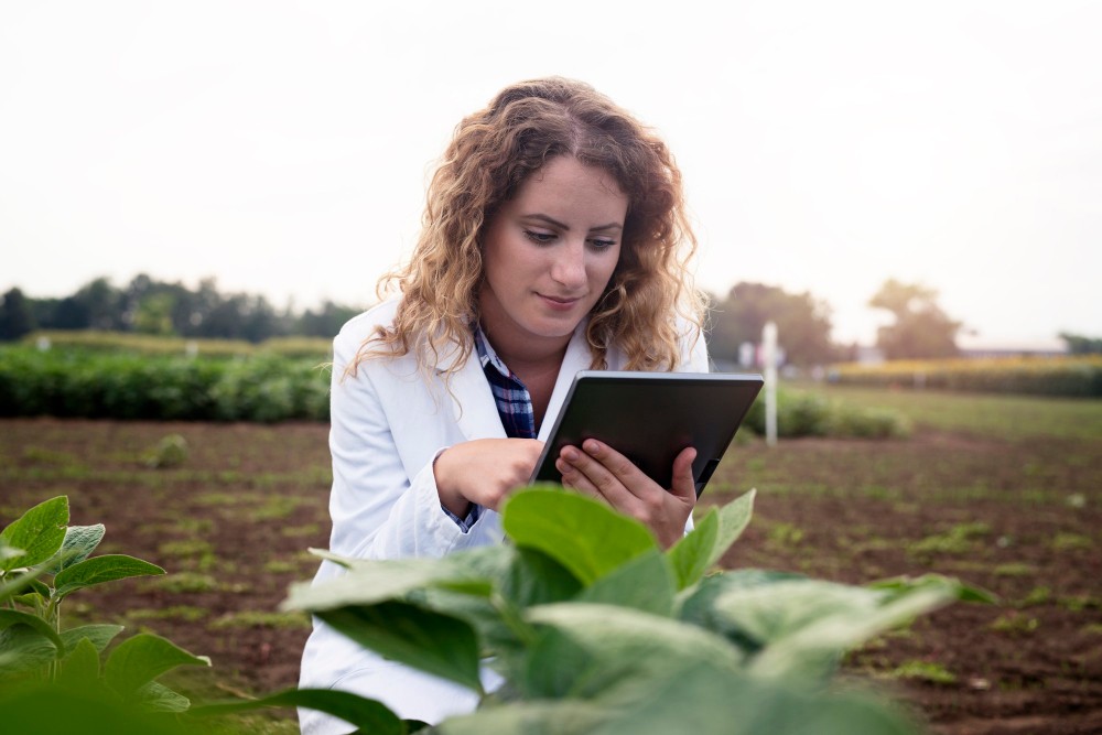 Mulher com jaleco em uma fazenda, examinando uma tela de tablet ao lado de plantas e cultura agrícola ao ar livre, ao entardecer.