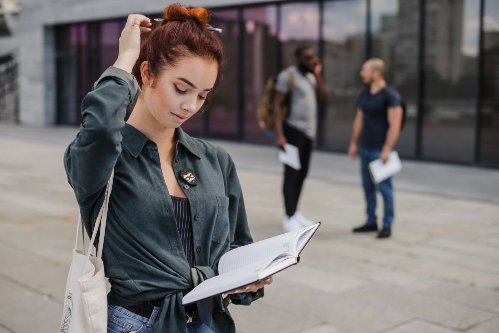 Mulher com cabelo ruivo lendo um caderno aberto na rua, usando jaqueta escura e bolsa de tecido, enquanto outras pessoas aparecem ao fundo em ambiente urbano