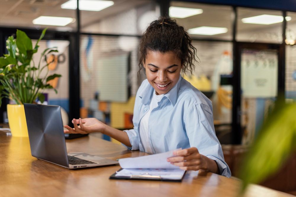 Mulher sorridente em escritório moderno, analisando documentos e usando notebook para trabalho, em ambiente de coworking com mesa e plantas ao fundo.