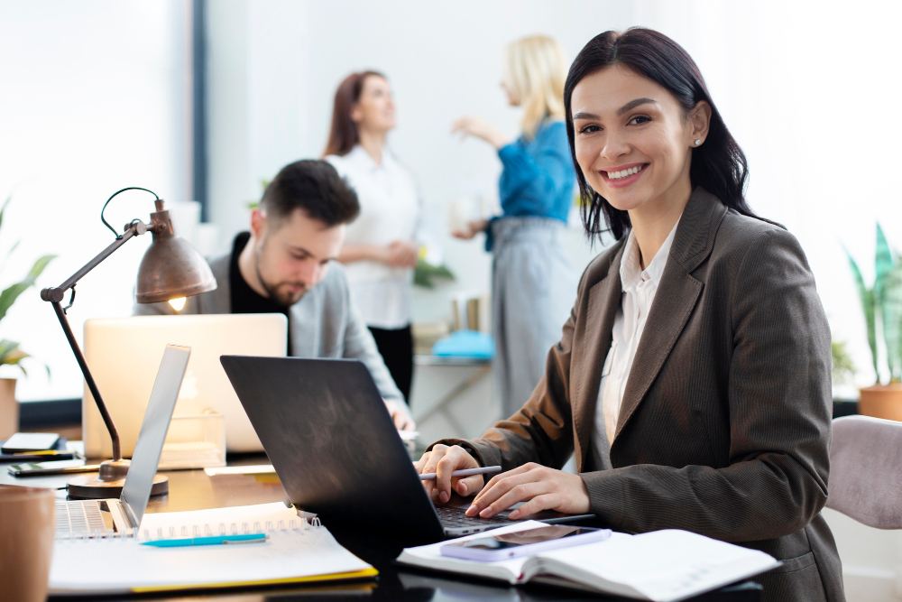 Mulher em ambiente corporativo sorrindo enquanto trabalha em um notebook no escritório, com colegas ao fundo usando computadores, iluminação suave e clima profissional.