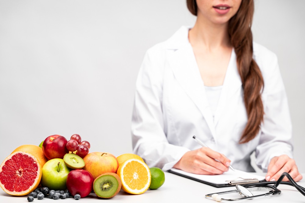 Nutricionista em jaleco branco analisando um bloco de anotações ao lado de frutas frescas como laranja, maçã, kiwi, banana e uvas, em ambiente clean