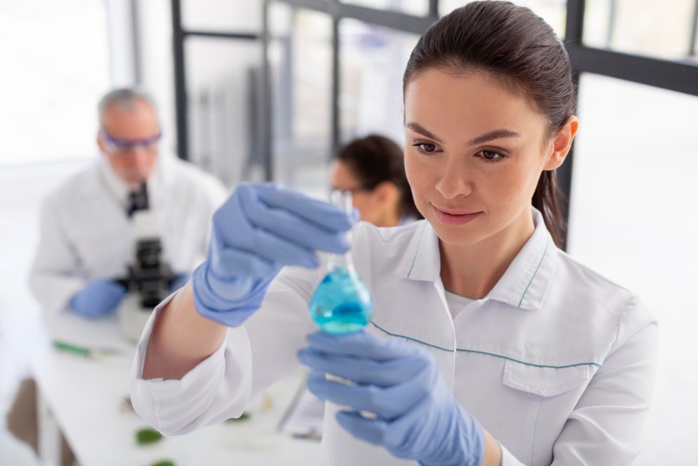 Pesquisadora em laboratório de ciências com luvas azuis, realizando manuseio cuidadoso de um tubo com solução azul enquanto colegas observam ao fundo.