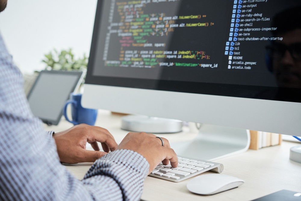 Pessoa trabalhando em computador com código de programação na tela, usando teclado e mouse em ambiente de escritório, com monitor e dispositivos sobre a mesa.