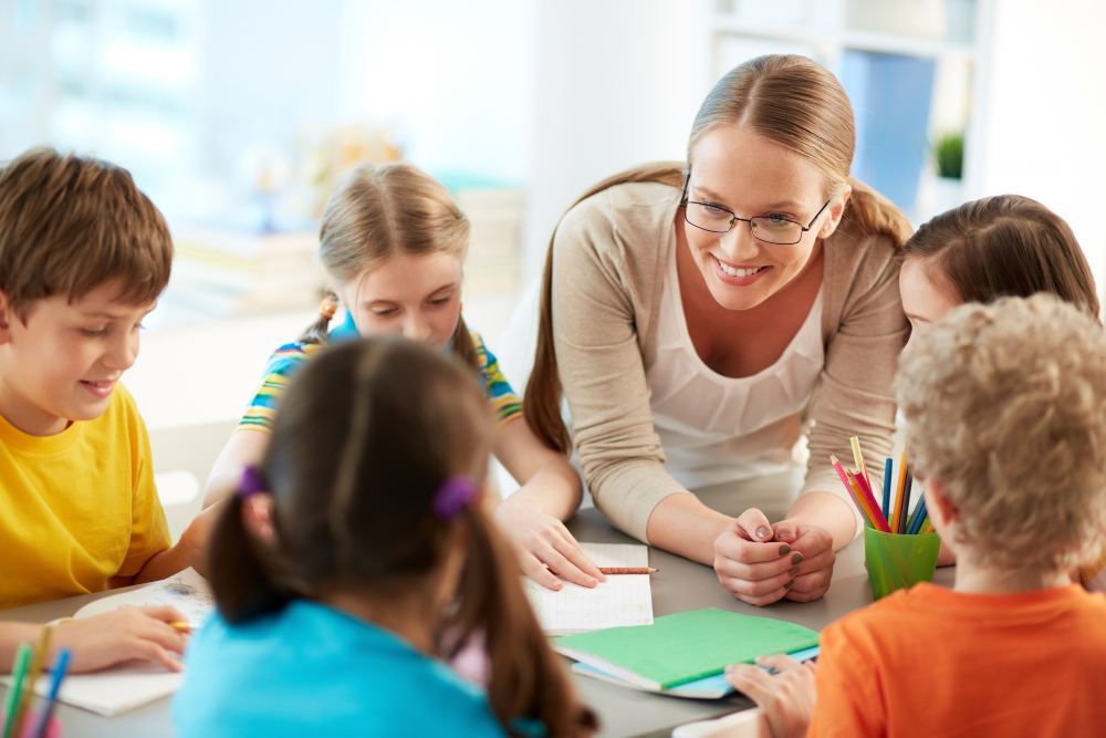 Professora sorridente auxilia alunos em sala de aula, ajudando crianças a desenhar e escrever em mesas com lápis de cor, cadernos e material escolar.