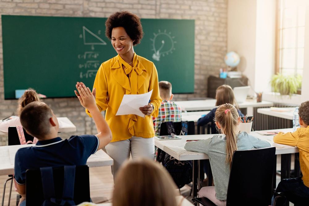 Professora em sala de aula com quadro verde, sorrindo e entregando uma folha enquanto alunos prestam atenção e levantam a mão, em aula de educação infantil.