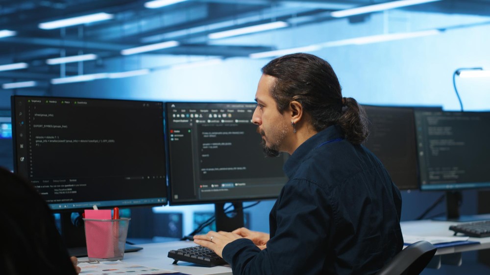 Profissional de tecnologia sentado em frente a monitores com código e linhas de programação em ambiente de escritório, olhando para a tela enquanto trabalha.