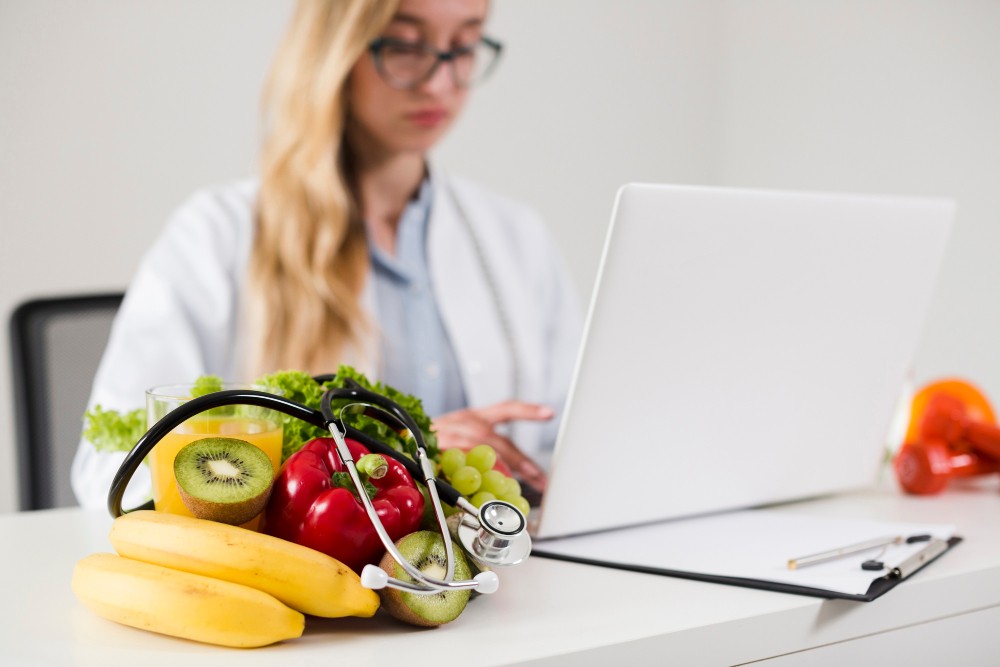 Profissional de saúde em escritório com estetoscópio sobre a mesa ao lado de frutas e legumes, com notebook aberto para consulta e planejamento alimentar.