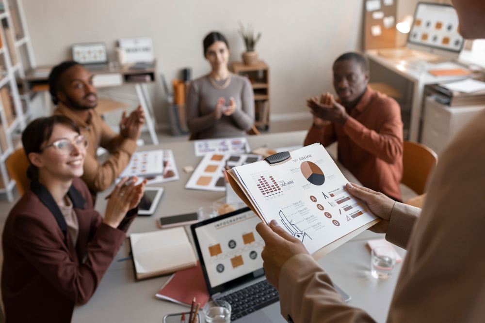 Reunião em equipe em escritório, com pessoas analisando gráficos e relatórios impressos em uma mesa durante uma apresentação de dados.