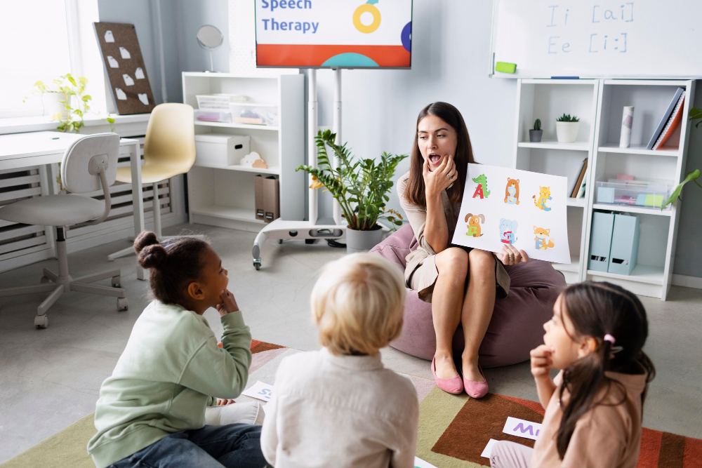 Sessão de terapia e aprendizado infantil em sala de aula: uma terapeuta sentada no chão mostra cartões educativos com letras para crianças sentadas ao redor, em ambiente organizado com mesas e prateleiras ao fundo.