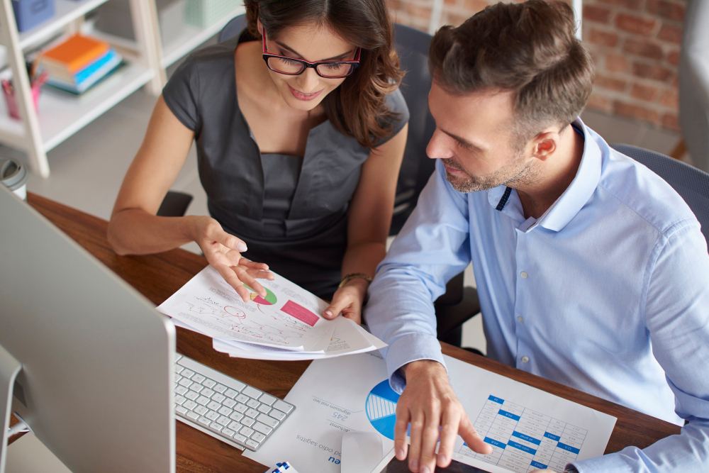 Profissional e equipe analisam relatórios financeiros e gráficos em um escritório moderno, apontando dados em uma planilha impressa sobre a mesa com notebook.
