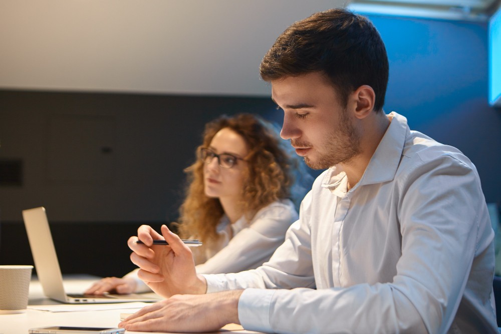 Homem e mulher trabalhando em equipe em um escritório moderno, com o homem analisando dados no laptop e a colega ao fundo.