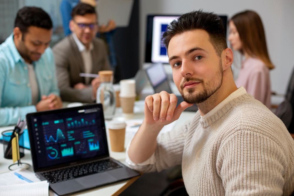 Homem jovem em escritório de tecnologia, com tela de computador e gráficos na mesa ao fundo, enquanto conversa e observa com expressão pensativa durante reunião