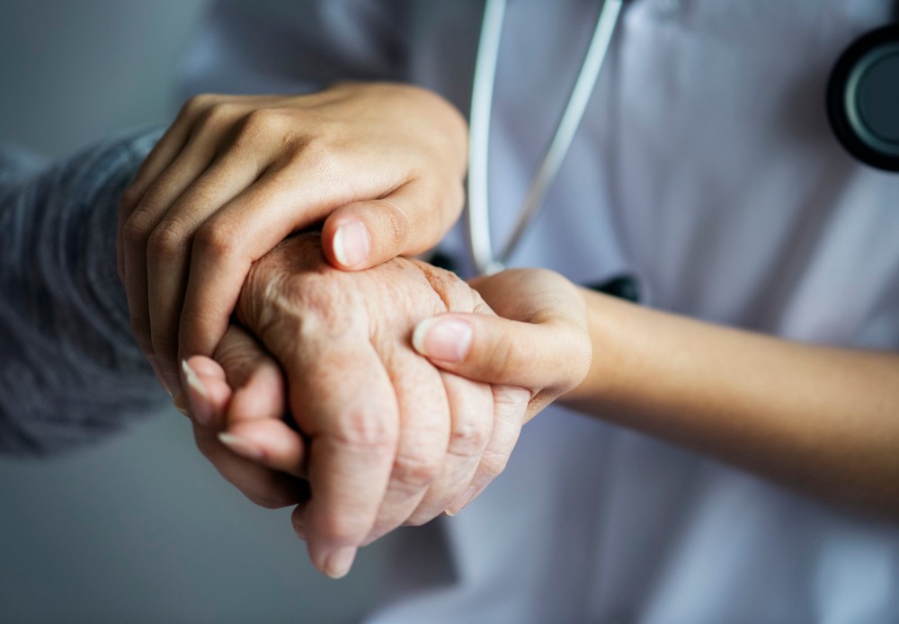 Médico segurando a mão de um idoso, gesto de cuidado e apoio durante atendimento de saúde, com foco nas mãos e na expressão de confiança.