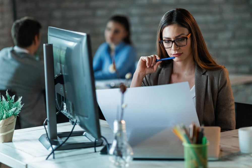 Mulher profissional com óculos usando fone de ouvido e concentrada em atendimento no computador em um escritório moderno, com documentos na mesa e colega ao fundo.