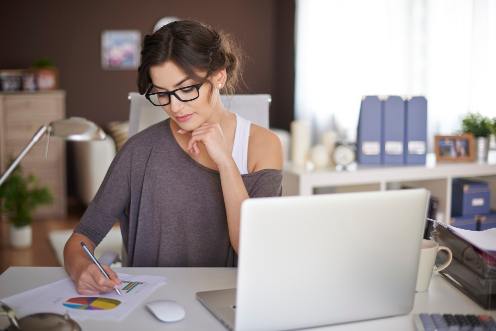 Mulher com óculos, em home office, escrevendo e desenhando ao lado de um notebook em uma mesa com papéis e gráficos coloridos.