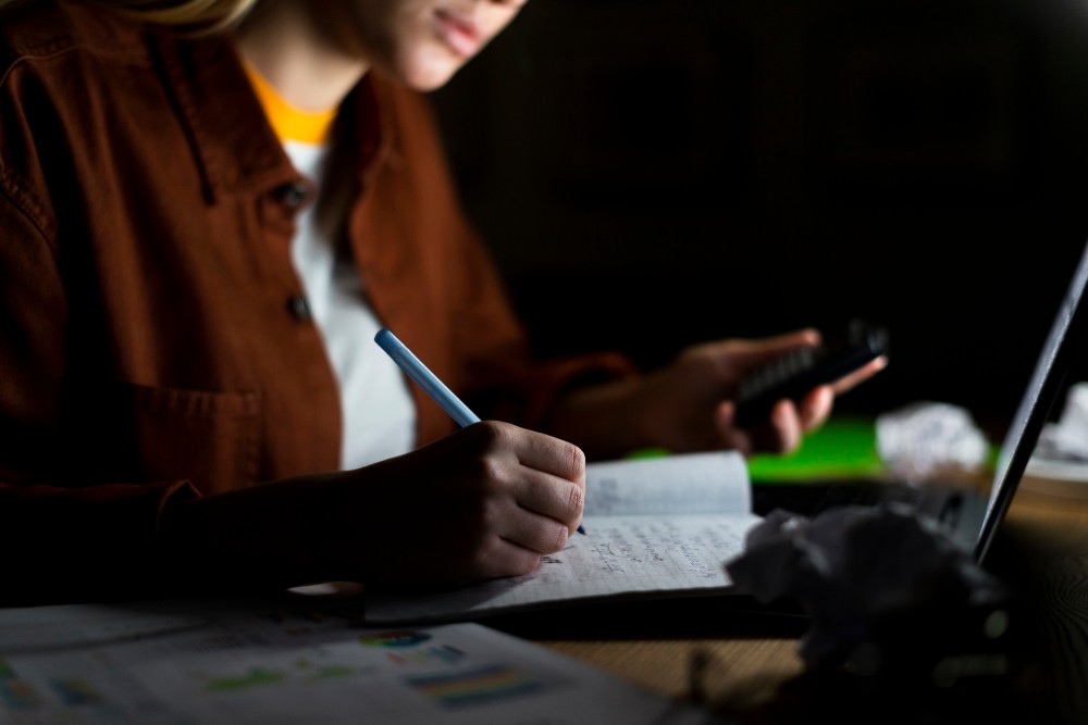 Pessoa escrevendo em um caderno com uma caneta azul, sentada diante de um notebook, em ambiente de estudo ou trabalho com foco no manuscrito.