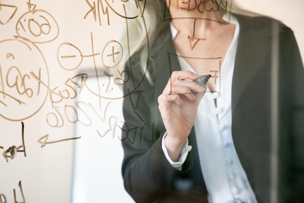 Pessoa em traje segurando um cartão eletrônico com anotações e fórmulas desenhadas em um quadro branco ao fundo, sugerindo planejamento e tecnologia