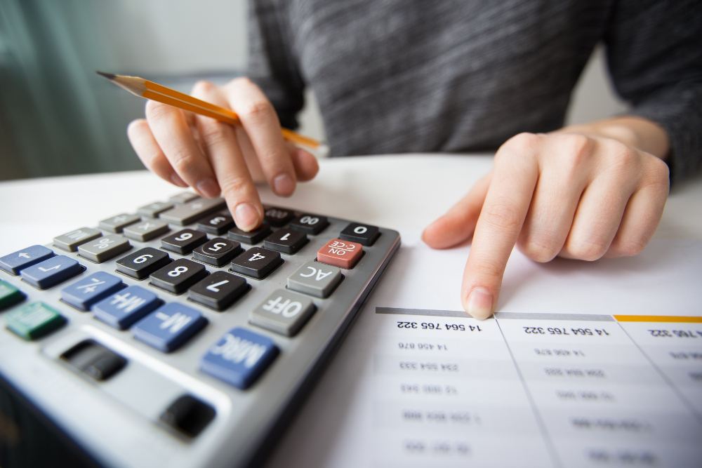 Pessoa usando calculadora e revisando planilhas financeiras em uma mesa, com anotações e documentos ao lado para conferência de números.