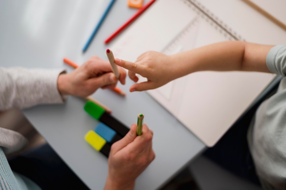 Pessoas em uma reunião de trabalho apontam e colocam lápis sobre cartões coloridos e um quadro branco, sugerindo planejamento, organização e colaboração em equipe.