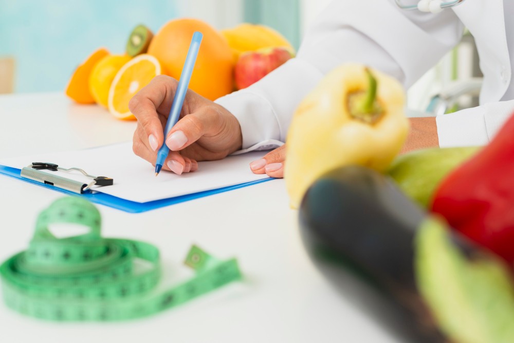 Nutricionista ou profissional de saúde registra informações em prancheta enquanto avalia alimentos, como pimentões e frutas, em ambiente clínico ou de atendimento.