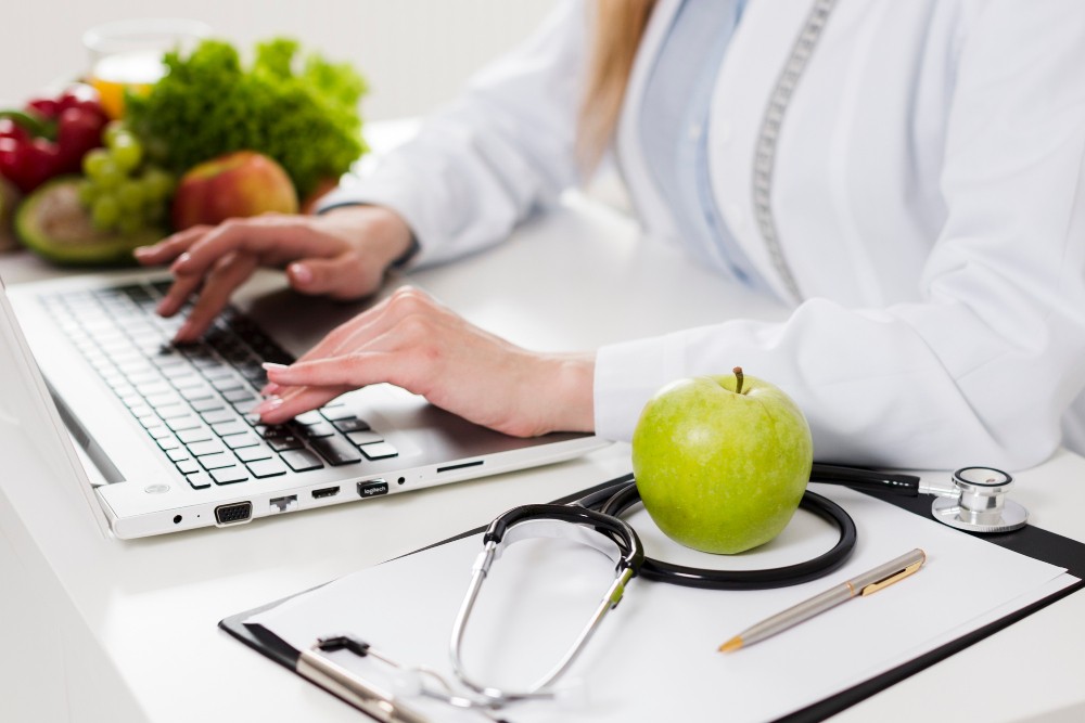 Profissional de saúde digitando em notebook no escritório, com maçã verde, estetoscópio e prancheta sobre a mesa, sugerindo alimentação e bem-estar