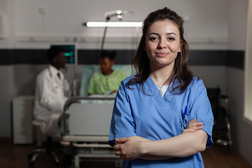 Profissional de saúde em pé com braços cruzados, usando uniforme azul, em ambiente hospitalar com leitos ao fundo e equipe desfocada